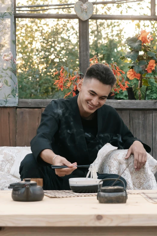 a man sitting on top of a couch next to a table, a portrait, inspired by Itō Ogura Yonesuke, trending on unsplash, people outside eating meals, wearing black silk robes, joe keery, smiling playfully