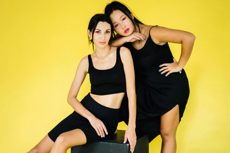 a couple of women standing next to each other, inspired by Wang Duo, trending on pexels, wearing a cropped black tank top, sitting pose, yellow, wearing a black dress