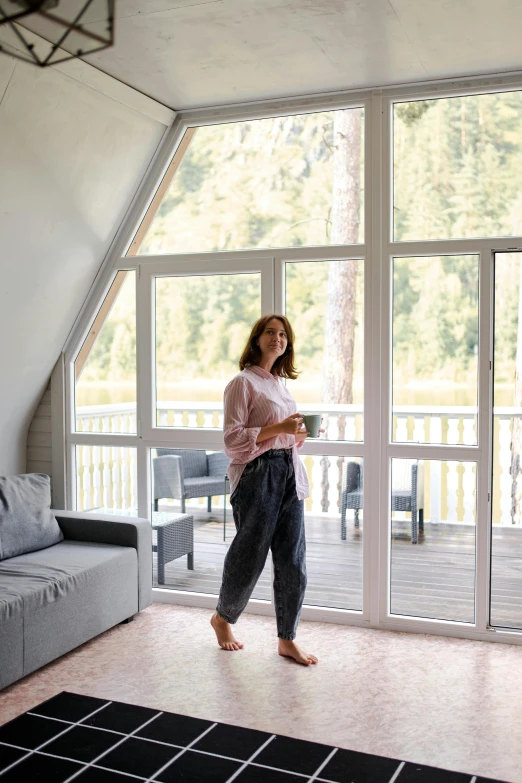 a woman standing in a living room next to a couch, inspired by Constantin Hansen, luxurious wooden cottage, balcony, glamping, grey