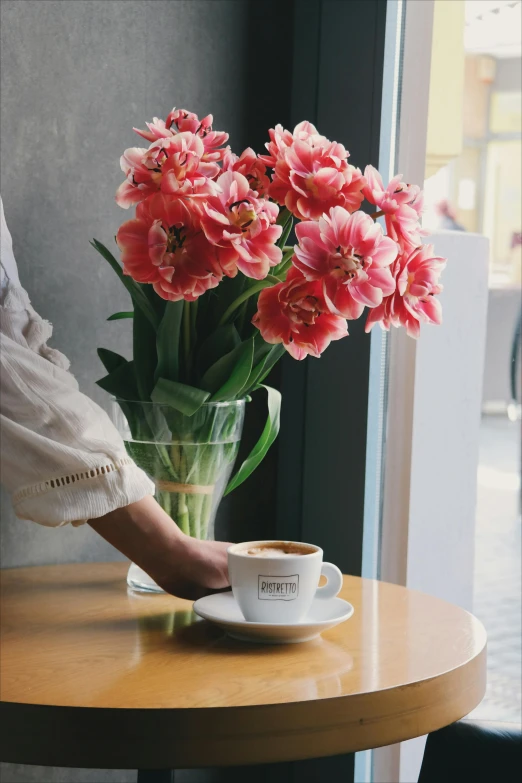 a person holding a cup of coffee near a vase of flowers, inspired by Louis Stettner, at the counter, city views, tulip, pink flowers