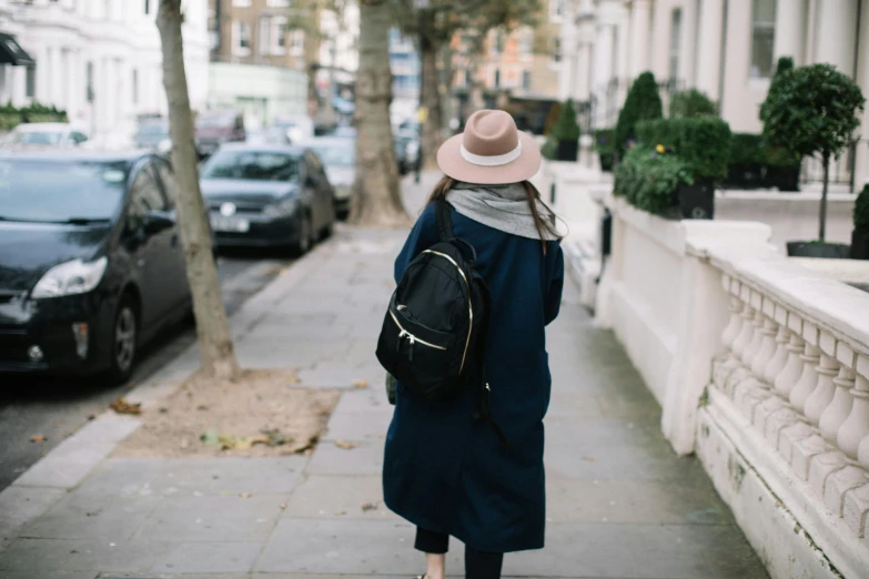 a woman walking down a sidewalk with a backpack, by Nina Hamnett, pexels contest winner, emma watson wearing fancy hat, blue coat, square backpack, unsplash photography