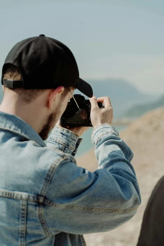 a man taking a picture of another man with a camera, trending on pexels, wearing a backwards baseball cap, mountains, wearing jeans and a black hoodie, looking down on the camera