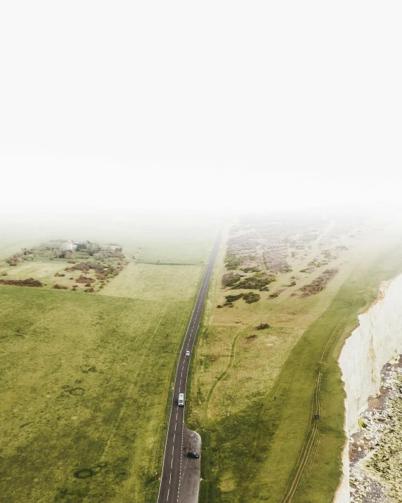 a car driving down a road next to a lush green field, an album cover, pexels contest winner, cliffs of dover, foggy photo 8 k, birds eye, boundary of two lands