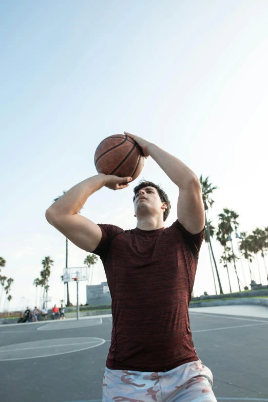 a man standing on top of a basketball court holding a basketball, trending on dribble, with palm trees in the back, shooting angle from below, wearing a muscle tee shirt, broad shoulder