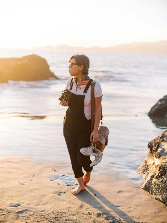 a woman standing on a beach holding a camera, wearing overalls, zenra taliyah, sunset magazine, profile image