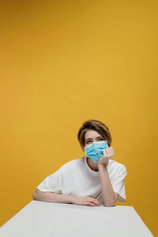 a person sitting at a table with a face mask on, a picture, trending on pexels, happening, yellow backdrop, cysts, mouth shut, blue