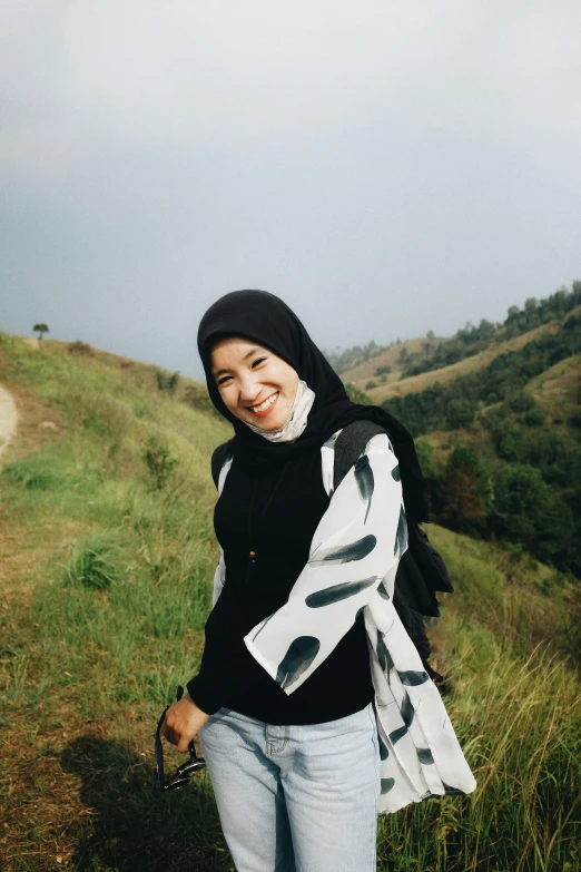 a woman standing on top of a lush green hillside, a picture, inspired by JoWOnder, sumatraism, white hijab, very slightly smiling, black in, low quality photo