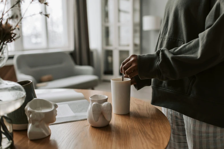 a person standing in a living room next to a table, a still life, trending on pexels, paper cup, pouring, white sleeves, porcelain organic