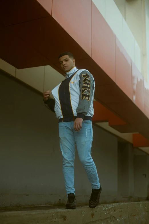 a man standing on a ledge in front of a building, an album cover, inspired by Kyle Lambert, trending on pexels, renaissance, bomber jacket, striking a pose, wearing jeans, professional sports style