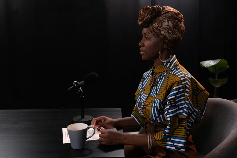 a woman sitting at a table with a cup of coffee, inspired by Carrie Mae Weems, afrofuturism, cloth head wrap, holding a microphone, production photo, octa 8k