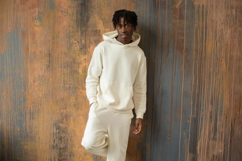 a man standing in front of a rusty wall, a picture, inspired by Thomas Blackshear, wearing a hoodie and sweatpants, ivory, pearlescent white, off-white