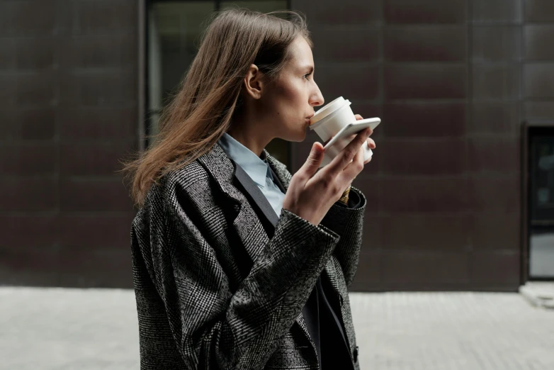 a woman is drinking out of a cup, by Emma Andijewska, trending on unsplash, wearing a worn out suit, wearing a long coat, trending on dezeen, exterior shot