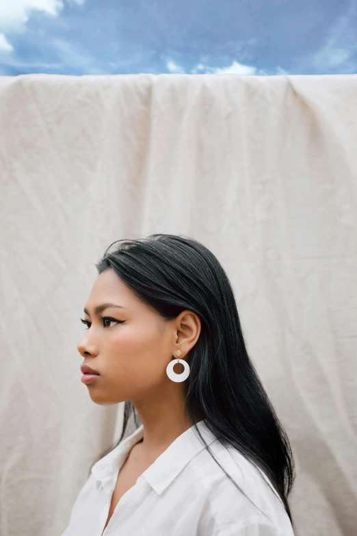 a woman standing in front of a white curtain, inspired by Ruth Jên, trending on unsplash, segmented 2d laser cut earrings, south east asian with round face, on vellum, woman in streetwear