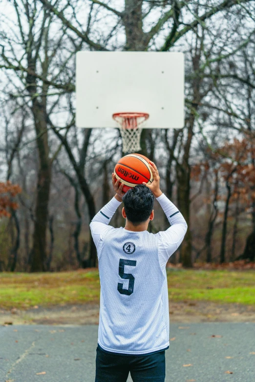 a man standing on top of a basketball court holding a basketball, a picture, by Greg Spalenka, trending on dribble, at a park, facing away from camera, white sleeves, against the backdrop of trees