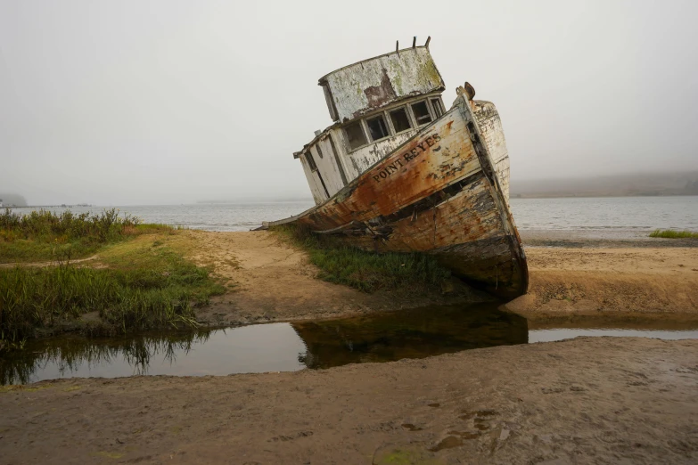 a boat sitting on top of a beach next to a body of water, by Jessie Algie, rusted silent hill, moist foggy, bay area, where a large