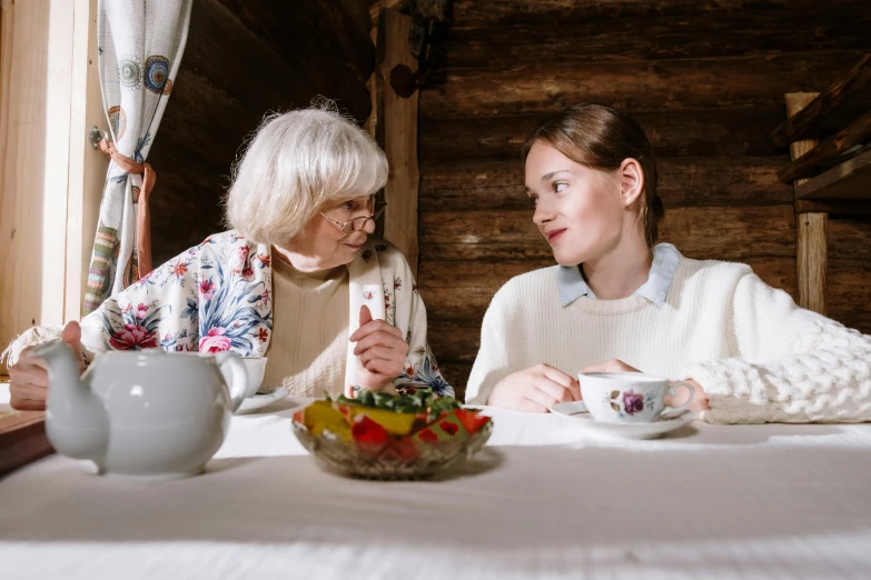 two women sitting at a table with a bowl of fruit, by Adam Marczyński, pexels contest winner, in a cabin, white haired lady, old and young, with a white mug