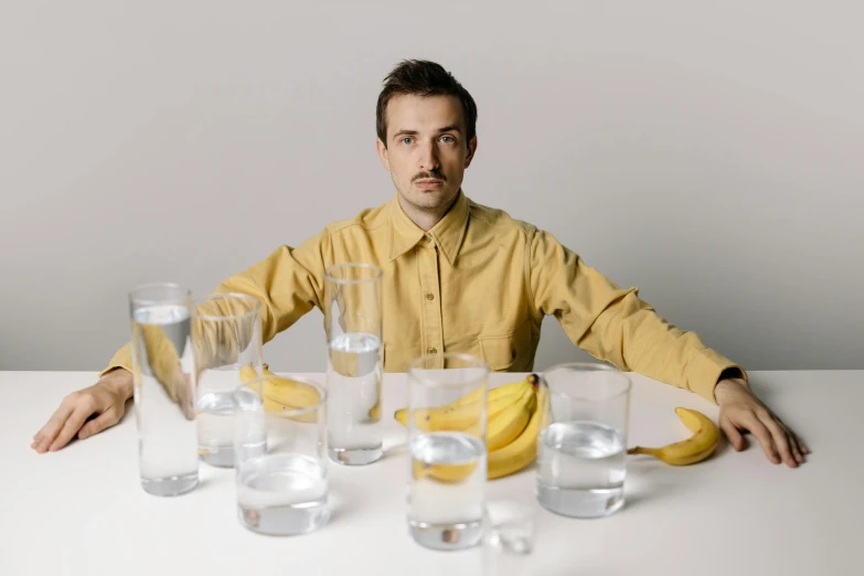 a man sitting at a table with glasses and a banana, water water, sydney hanson, on a pale background, press shot