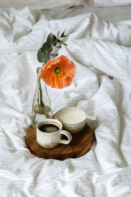 an orange flower sitting in a vase next to a cup of coffee, un made bed, on a wooden tray, profile pic, uncropped