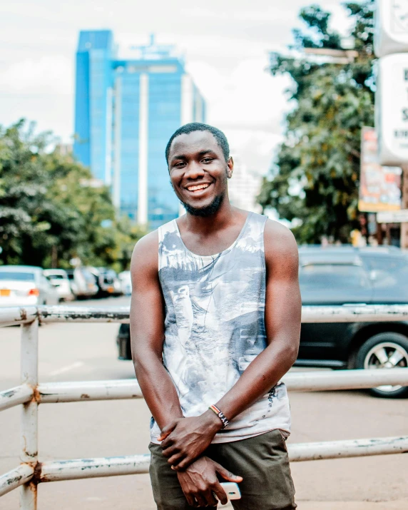 a man standing in front of a fence on a city street, by Ingrida Kadaka, pexels contest winner, happening, smiling sweetly, emmanuel shiru, with grey skin, 25 years old