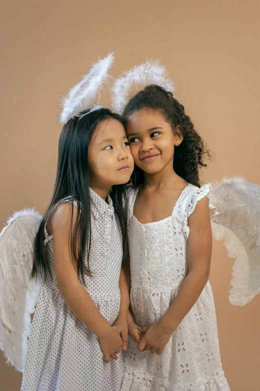 a couple of little girls standing next to each other, by Marie Angel, pexels contest winner, symbolism, large white wings, varying ethnicities, studio photo, mai anh tran