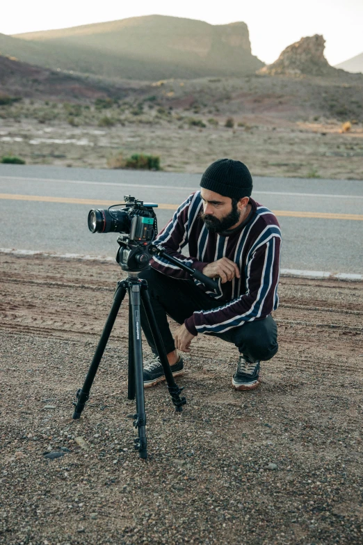 a man kneeling down next to a camera on a tripod, a picture, unsplash contest winner, video art, screenwriter, a portrait of rahul kohli, desert photography, grainy footage