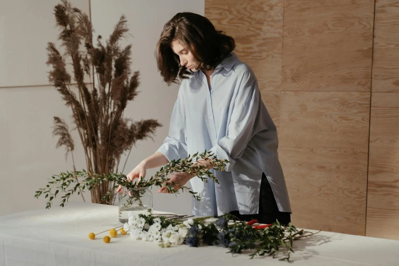 a woman arranging flowers in a vase on a table, by Alice Mason, trending on unsplash, wearing a white button up shirt, white sweeping arches, backdrop, showpiece