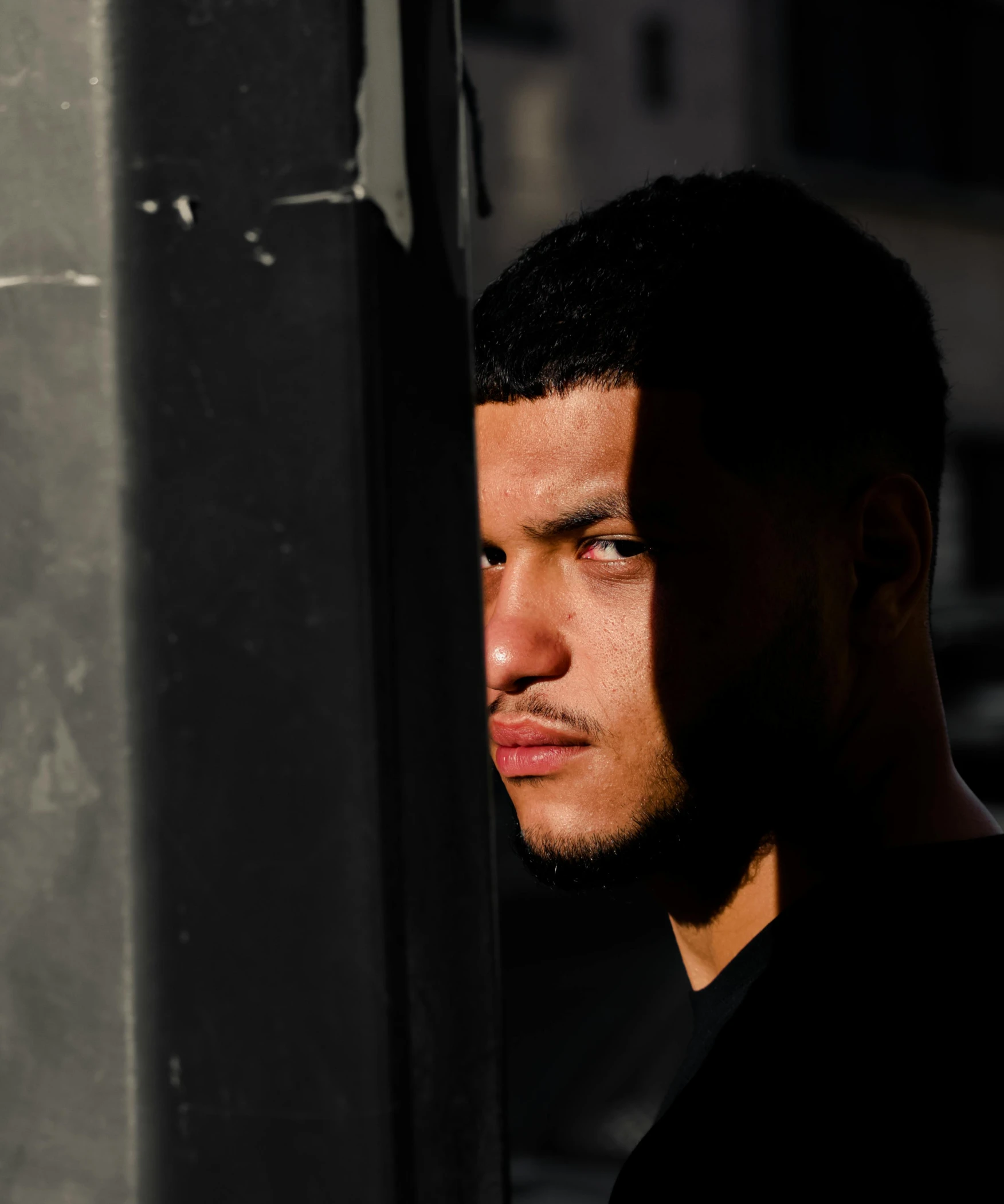 a man in a black shirt leaning against a pole, pexels contest winner, light-brown skin, face in the shadows, joseph moncada, backlit ears