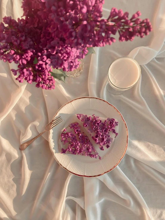 a white plate topped with a piece of cake next to a purple flower, a picture, by Lucia Peka, diaphanous iridescent cloth, gif, promo image, 5k