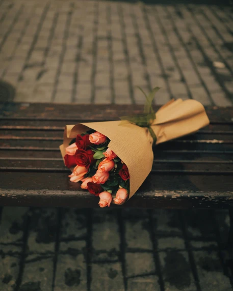 a bunch of flowers sitting on top of a wooden bench, holding a rose, on the sidewalk, wrapped, dark. no text
