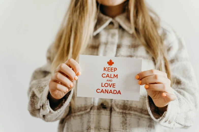 a woman holding a sign that says keep calm and love canada, by Julia Pishtar, pexels contest winner, american postcard art style, white, flannel, b