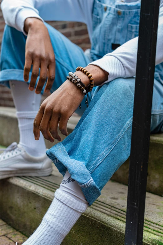 a person that is sitting on some steps, wearing two silver bracelets, wearing double denim, brown clothes, kano)