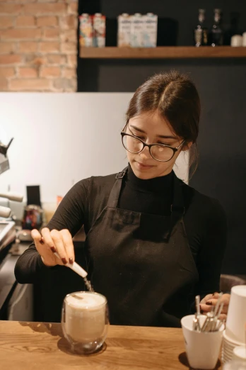a woman making a drink in a coffee shop, pexels contest winner, mia khalifa, aussie baristas, wearing black frame glasses, wearing an apron