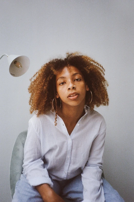 a woman sitting on top of a chair next to a lamp, by Lily Delissa Joseph, trending on unsplash, renaissance, natural hair, looking straight to camera, on a white table, androgynous person