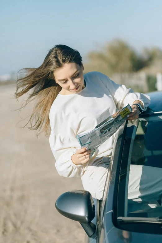 a woman leaning out the window of a car, reading the newspaper, wearing a white sweater, long hair windy, off-roading
