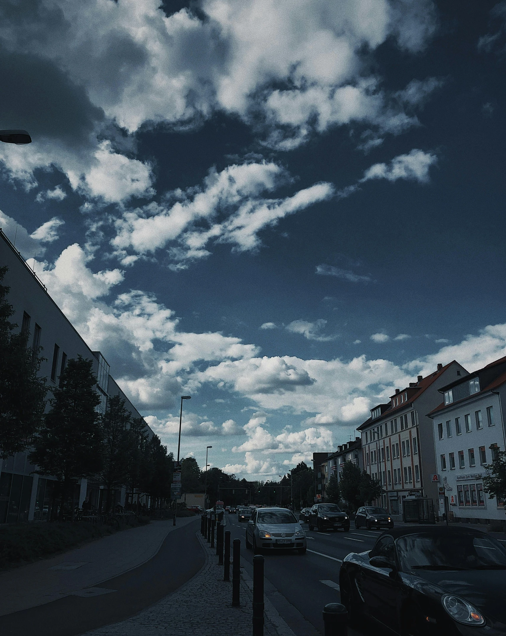 a street filled with lots of traffic next to tall buildings, a black and white photo, by Anna Haifisch, unsplash, beautiful sky with cumulus couds, a ghetto in germany, dark blue sky, low quality footage