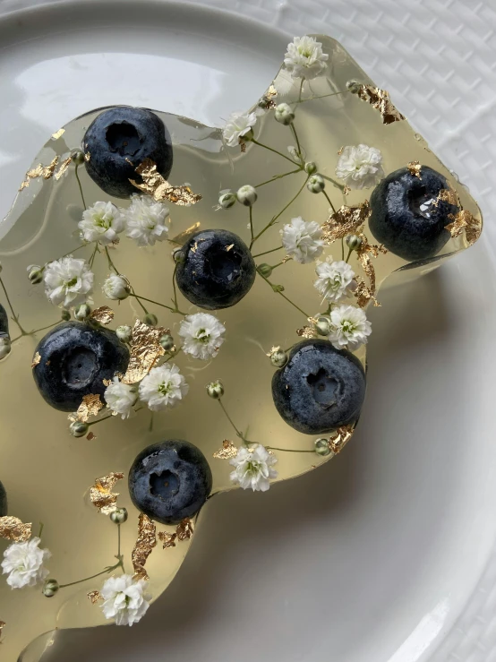 a white plate topped with blueberries and flowers, inspired by Barthélemy Menn, process art, stone and glass and gold, carved soap, unrefined sparkling gold nugget, high quality photo