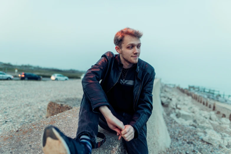 a man sitting on top of a rock next to the ocean, an album cover, by Thomas Furlong, unsplash, graffiti, wearing a leather jacket, slightly smiling, joel fletcher, soft light from the side