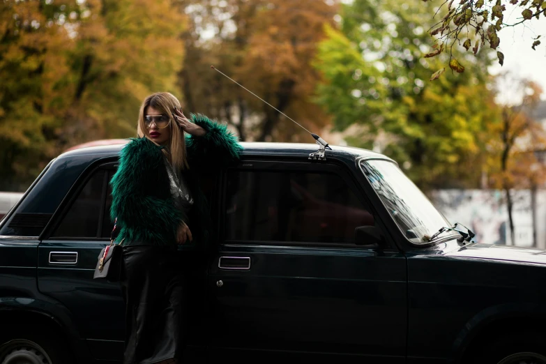 a woman leaning on the door of a black car, an album cover, inspired by Elsa Bleda, trending on pexels, realism, green fur, russian lada car, london fashion week, 15081959 21121991 01012000 4k