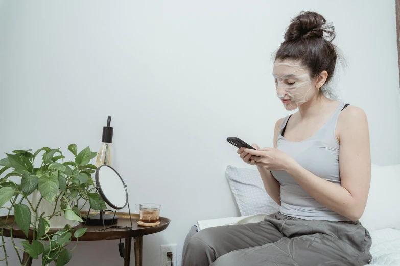 a woman sitting on a bed looking at her cell phone, by Adam Marczyński, trending on pexels, happening, wearing transparent glass mask, skincare, eucalyptus, gordon freeman as a woman