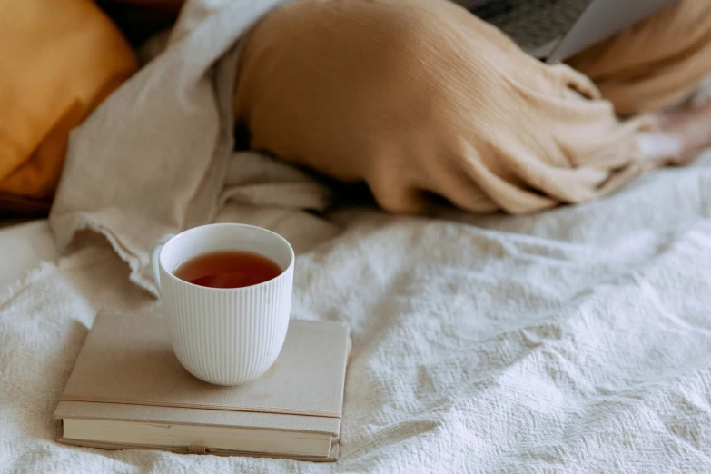 a person sitting on a bed with a book and a cup of tea, a still life, by Carey Morris, trending on pexels, linen, manuka, very crisp details, with a drink