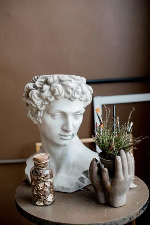 a statue sitting on top of a table next to a potted plant, trending on pexels, mannerism, handsome face, art supplies, white marble interior photograph, paining