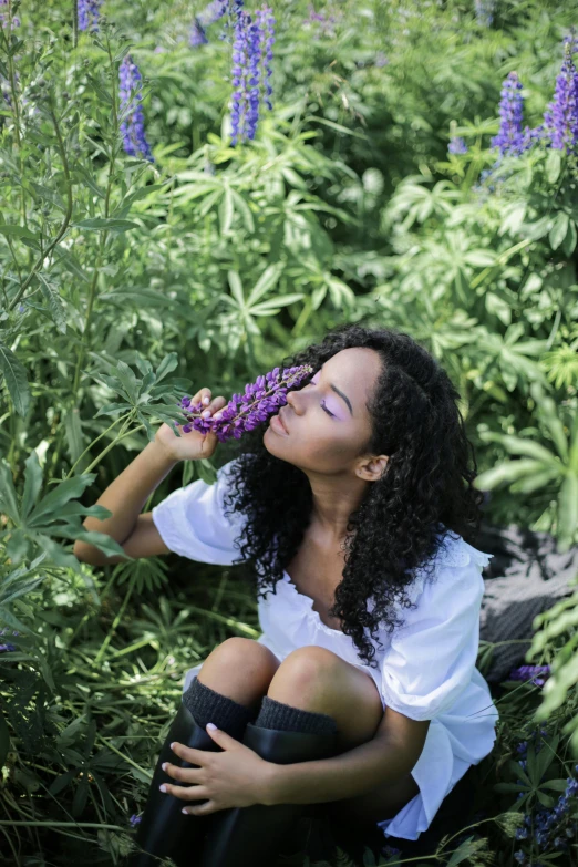 a woman sitting in a field of purple flowers, smelling good, with long curly, next to a plant, ashteroth