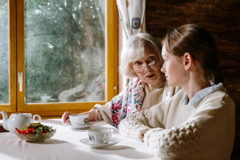 two women sitting at a table in front of a window, by Emma Andijewska, pexels contest winner, an old lady, in a cabin, avatar image, bashful expression