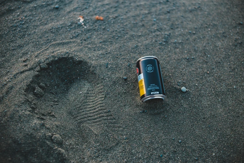 a can of beer sitting on top of a sandy beach, unsplash, hyperrealism, footprints, crashed in the ground, black sand, promo image
