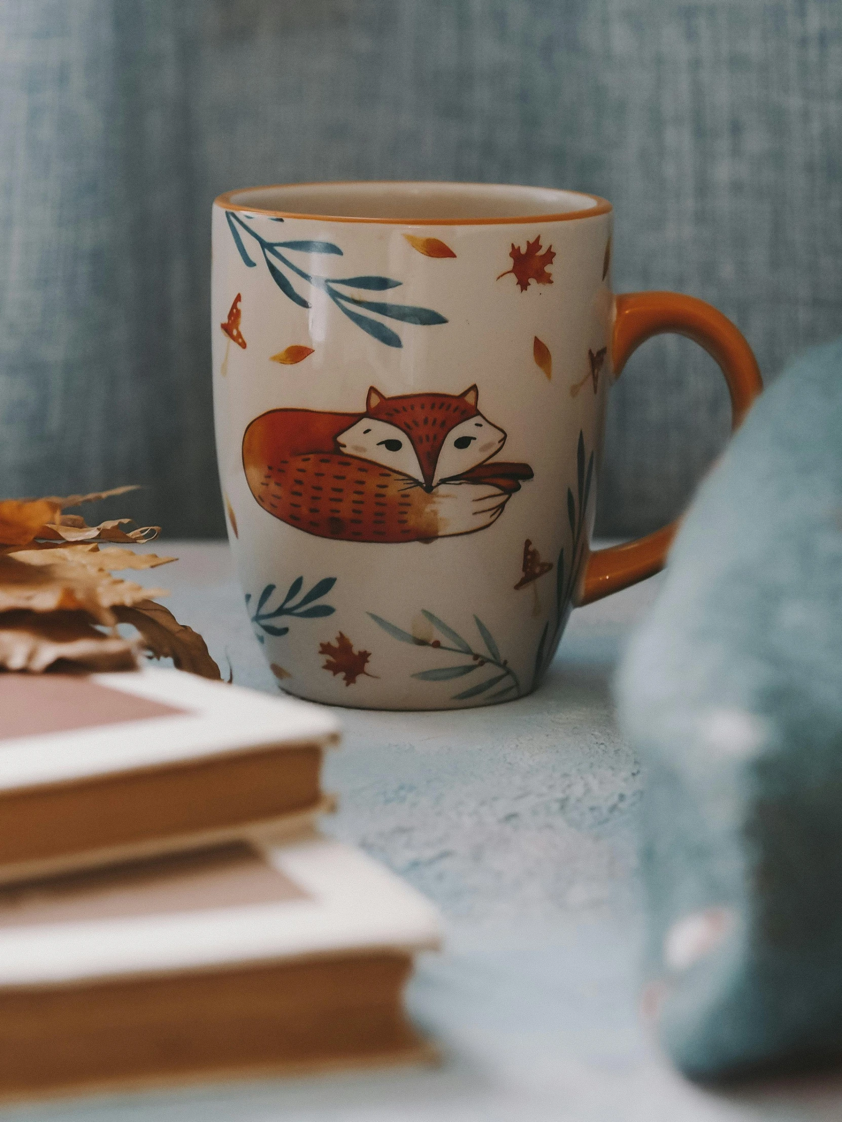 a coffee cup sitting on top of a table next to a pile of books, a still life, trending on pexels, folk art, cute fox, autumnal colours, detailed product image, front side view