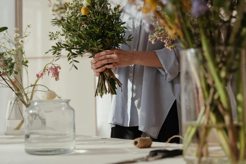 a woman holding a bunch of flowers in a vase, a still life, trending on unsplash, wearing a linen shirt, ingredients on the table, opening shot, herbs