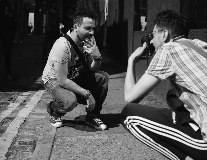 a couple of men sitting next to each other on a sidewalk, a black and white photo, happening, taking a picture, in an action pose, talking, adi meyers