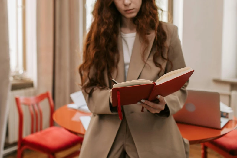 a woman standing in a room holding a book, trending on pexels, red jacket, curled up on a book, trench coat and suit, readhead