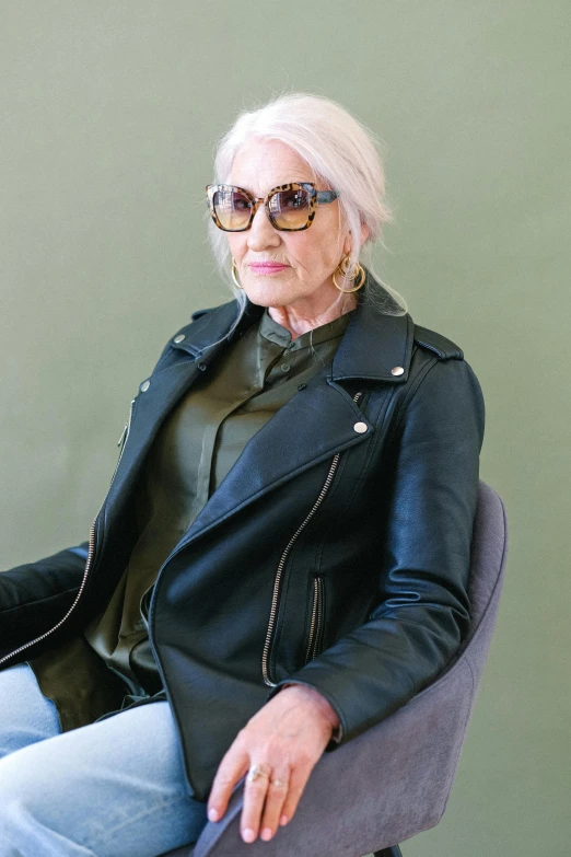 a woman sitting in a chair wearing a leather jacket, green shades, white haired, implanted sunglasses, profile image