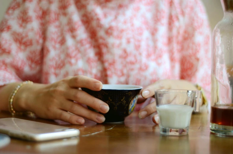 a woman sitting at a table with a cup of coffee, by Alice Mason, pexels, glass of milk, sake, hand on table, yogurt